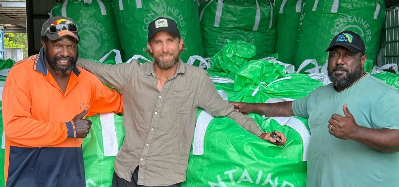 Two Crystal Ailan workers and a visitor standing in front of a wall of filled green Containers for Change collection bags at the Badu depot.