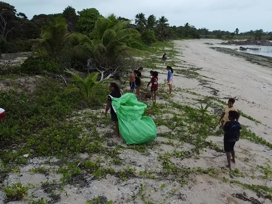 Community members walking a Badu Island shoreline during a clean-up, carrying a large green collection bag along the tide line.